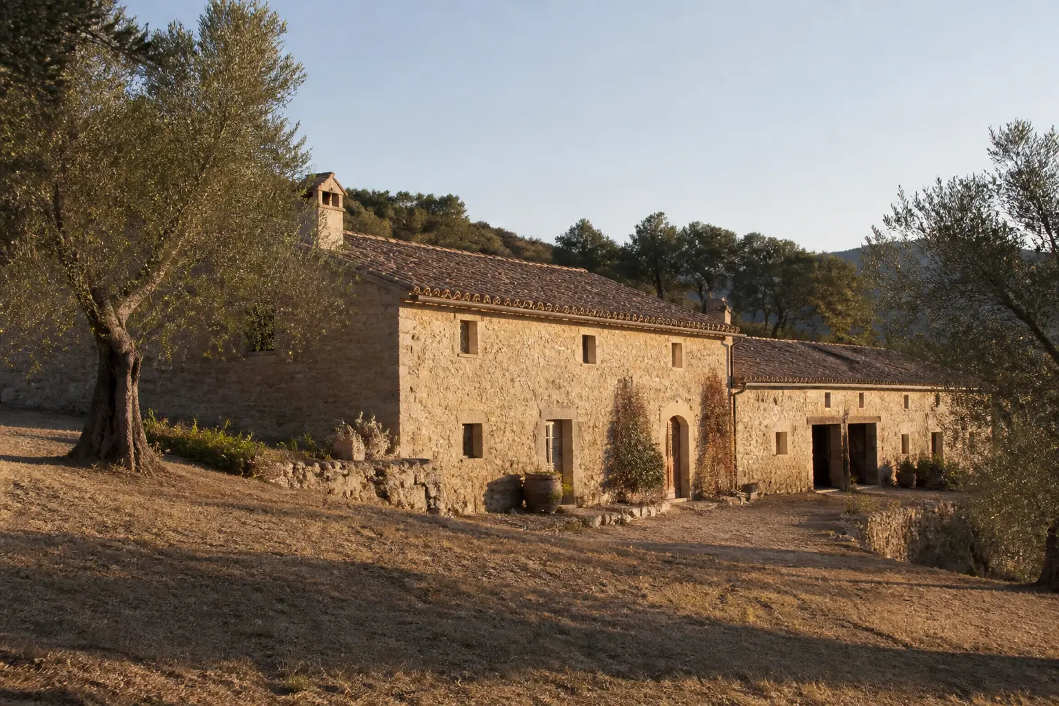 Maison provencale en pierre, long corps de ferme provençal sur terrain en pente, cour et oliviers au soleil couchant